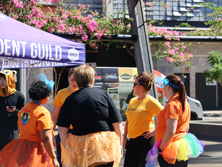 A group of people standing around a gazebo tent. The people are in bright fairy skirts and have colourful shirts on with them.