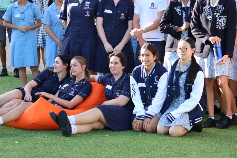 A photo of five high school students seated on the ground in front of more students. Three students are seated or kneeling on the lawn, while two students are lounging on a bright orange bean bag. 