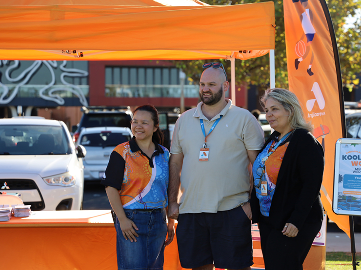 Three Anglicare WA employees standing in front of a Anglicare WA stall at a community event smiling at something out of shot.