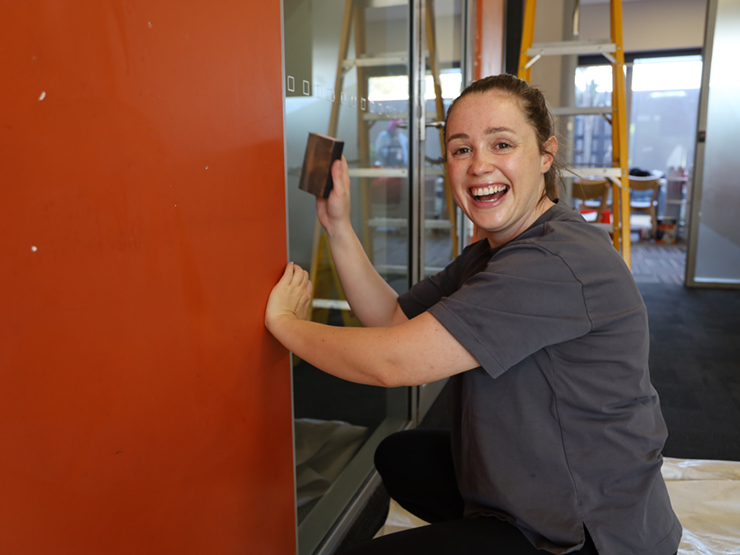 A person who is smiling at the camera while sanding down an orange wall so that it can be painted.