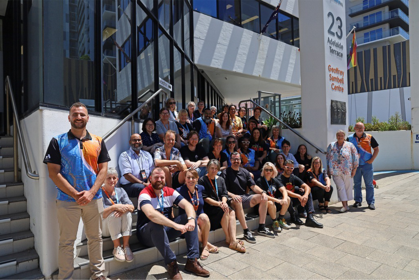 A large group of Anglicare WA staff sitting out the front of the Anglicare WA office building smiling at the camera.