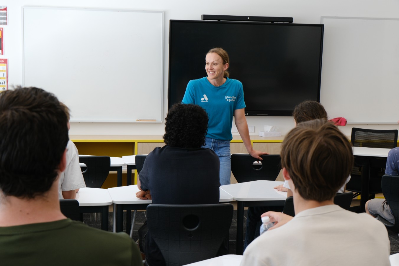 An educator wearing an Anglicare WA branded t-shirt smiling and speaking to a classroom of high school students. 