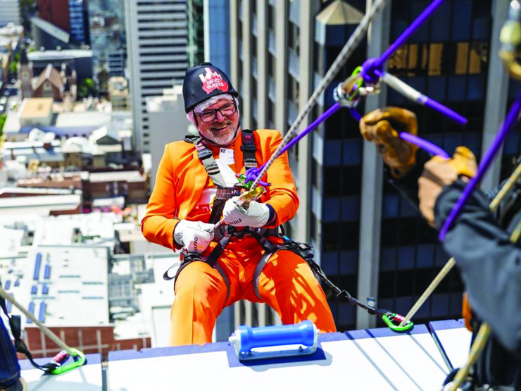 CEO Mark Glasson in an orange suit abseiling off the side of a building in the CBD. Mark is smiling at the camera.
