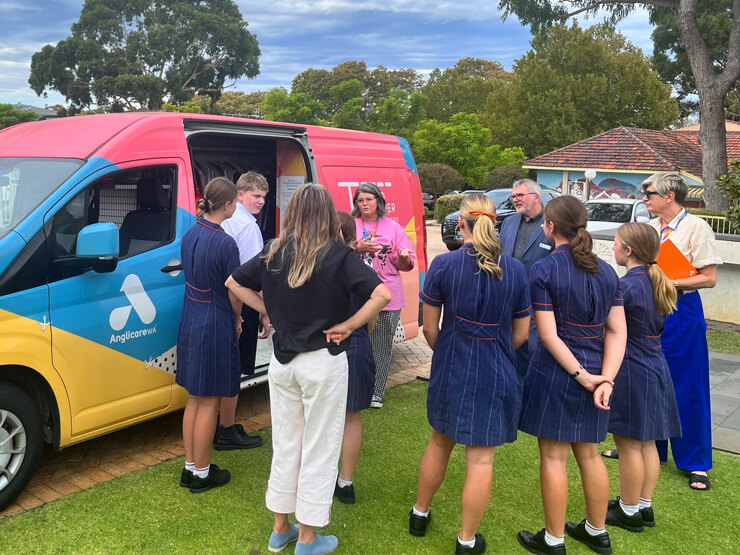 A photo of a group of adults and high school students standing beside a colourful van listening to an educator in a bright pink shirt speaking.  