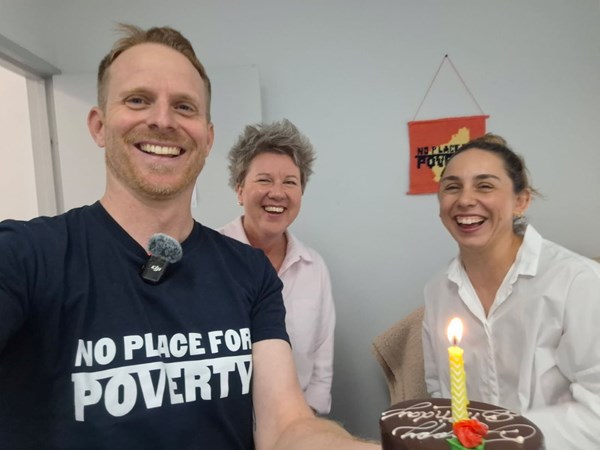 Three members of the No Place for Poverty team laugh while taking a selfie posing with a birthday cake topped with one candle.
