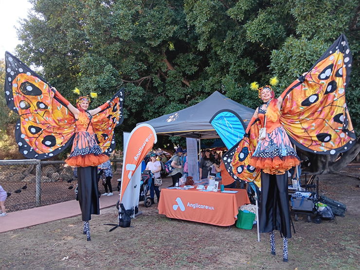 Two stilt walkers with butterfly wings standing in front of an Anglicare WA stall at a community event.