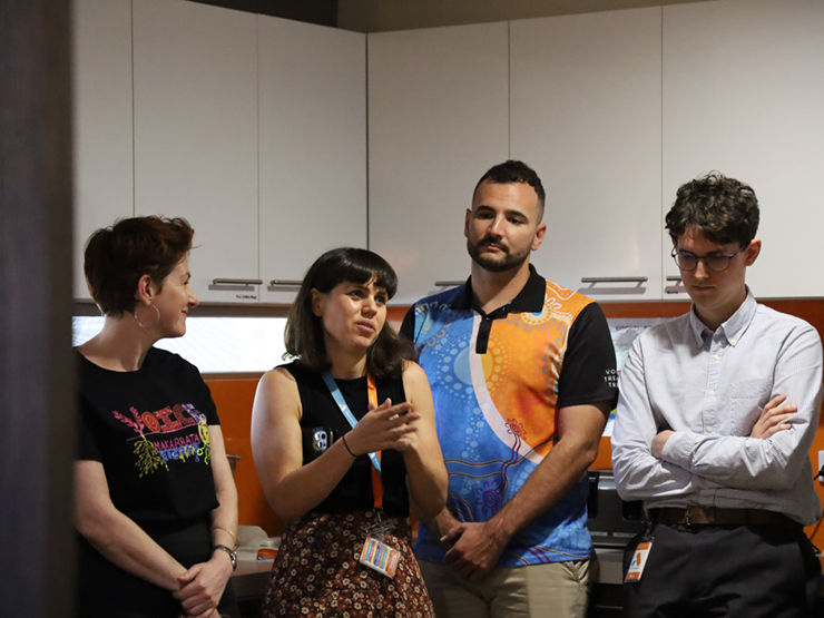 Four people standing next to each other with one of them talking to a wider group that are out of shot. The group are standing there listening the speaker intently.