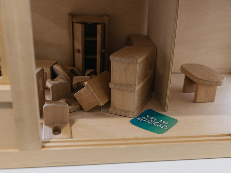 Close up shot through a window of a dolls house with furniture messily placed and a card that says 'you are making a difference'