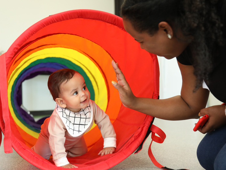 Toddler crawling through a plastic rainbow tunnel to a lady who is smiling and high fiving them.