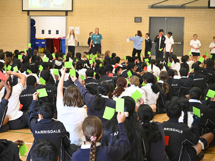 A photo of a school activity in a school gymnasium where many students sit cross legged on the floor holding up brightly coloured sticky note papers in the air.