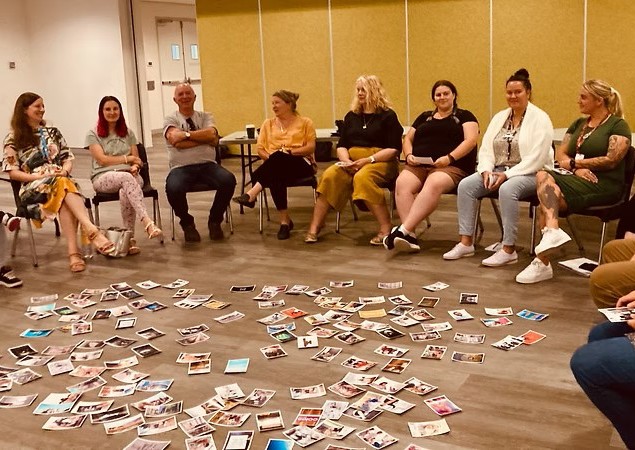 A photo taken during an ideation session of a group of people seated on chairs forming a circle with a collection of photographs strewn across the floor in the centre.