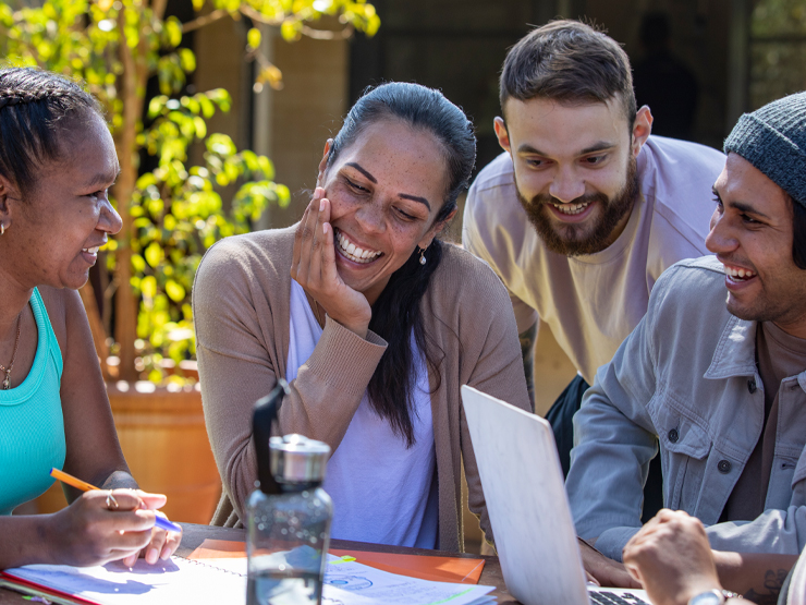 A group of young people gathered around a table, engaged with a laptop in a collaborative discussion