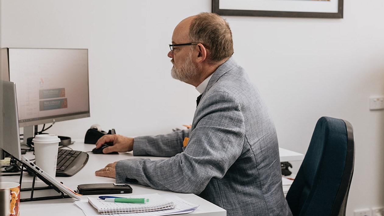 Anglicare WA Chaplain Chris Thomason in a grey blazer, seated at his desk working on the computer.
