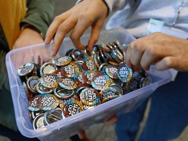 A plastic tub full on badges that have 'School Sleep Out' written on them, there are several hand reaching into the plastic tub pulling out badges.