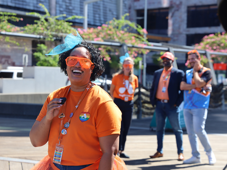 A Anglicare WA employee standing looking at the camera laughing. They are wearing an orange shirt, with orange novelty sunglasses on and a blue ribbon in their hair.