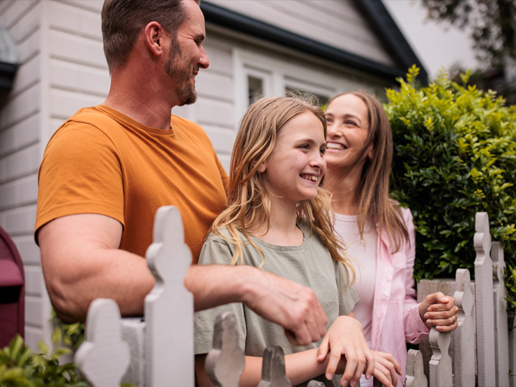 A family of four stands together in front of a white picket fence, smiling and enjoying a sunny day outdoors