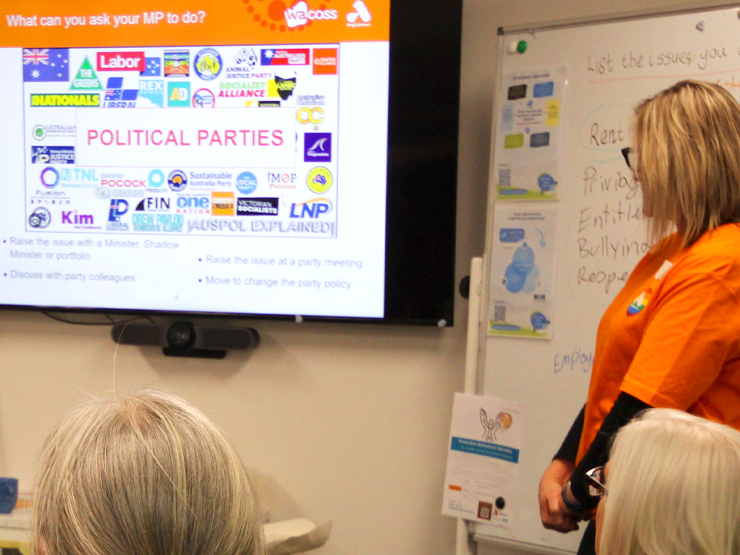 A Anglicare WA staff member standing next to a screen presenting to an audience. The screen has lots of different political party logo's on it. 