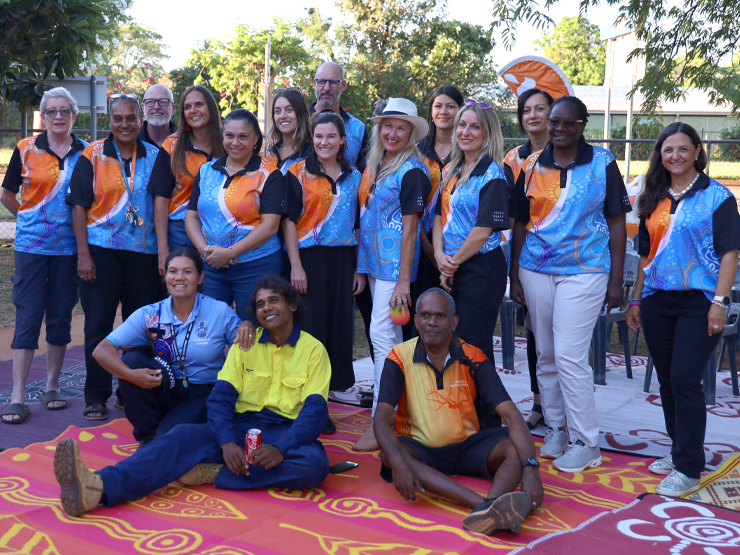 An image of a group of people who are standing looking at the camera and wearing matching blue and orange shirts with Australia Aboriginal artwork on them.