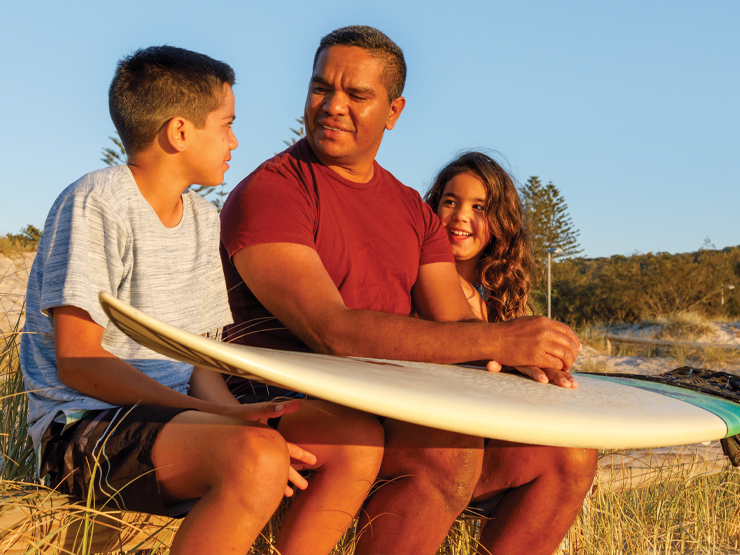 A man and two children are sitting together with a surfboard on their lap, enjoying a sunny day.