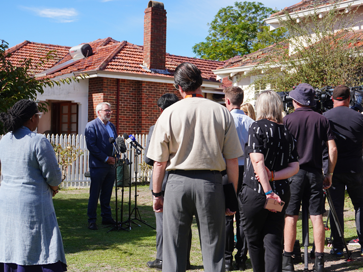 Mark Glasson CEO standing speaking to a group of media cameras in the front of a suburban house.
