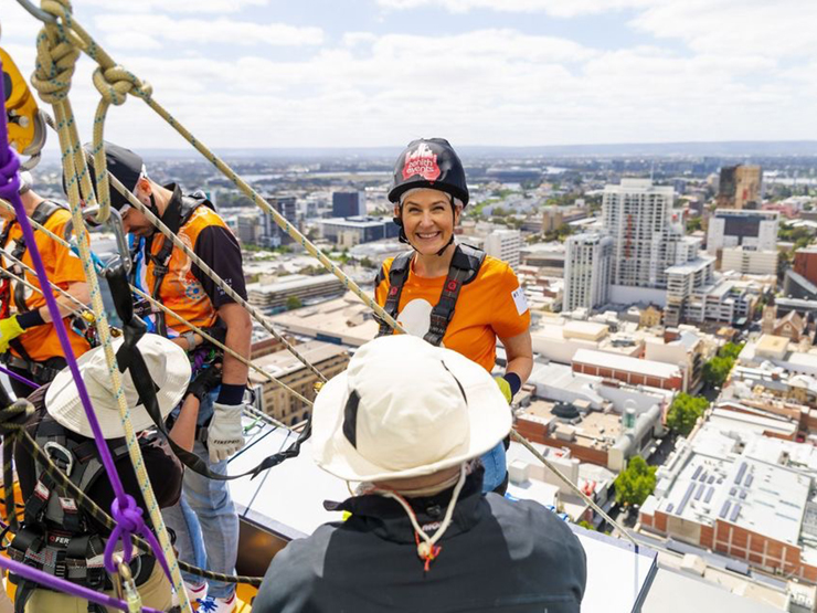A lady in a harness about to abseil off a building in the Perth CBD. The lady is wearing a orange shirt and is smiling at the camera.