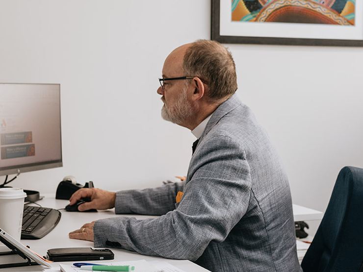 Image of a man wearing an Anglicare WA lanyard sitting looking at a computer screen working in an office.