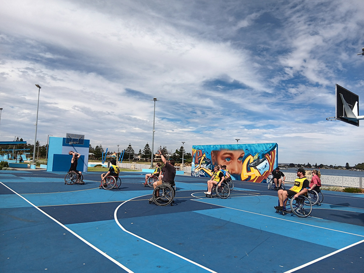 A seven people in wheelchairs playing basketball on an outside court on a sunny day.