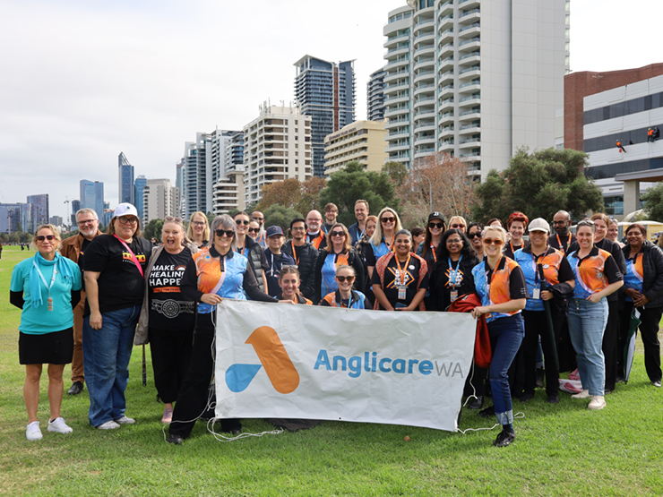 A large group of Anglicare WA staff looking at the camera at an event, they are carrying a Anglicare WA banner and smiling at the camera.