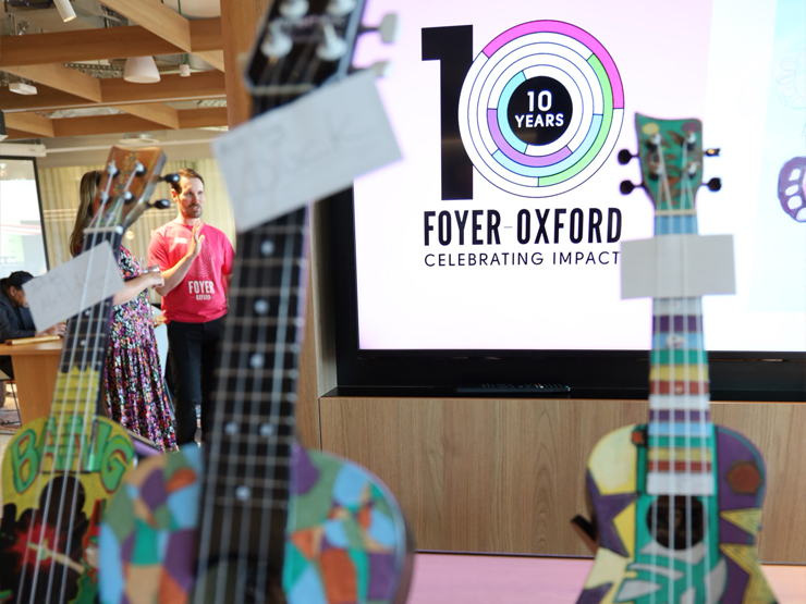 Close up of ukeleles which are out of focus, with a TV screen in the background that has 10 year of Foyer Oxford Celebrating Impact (this is a youth housing service run by Anglicare WA)