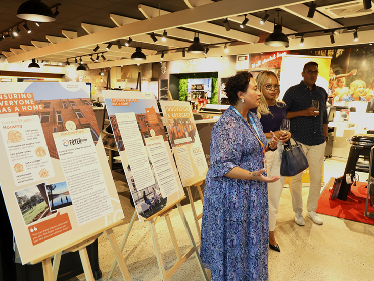 A female Anglicare WA talking to a group while standing next to 3 large boards with Anglicare WA advocacy areas on them.