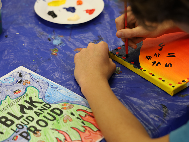 Image of child's hand painting on a small canvas at a table. Child is painting traditional Aboriginal motifs