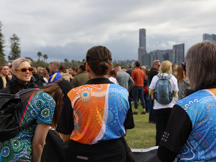 Image of the back of a group of people who are in a park at an event. They are wearing Anglicare WA shirts that have Aboriginal artwork on them.
