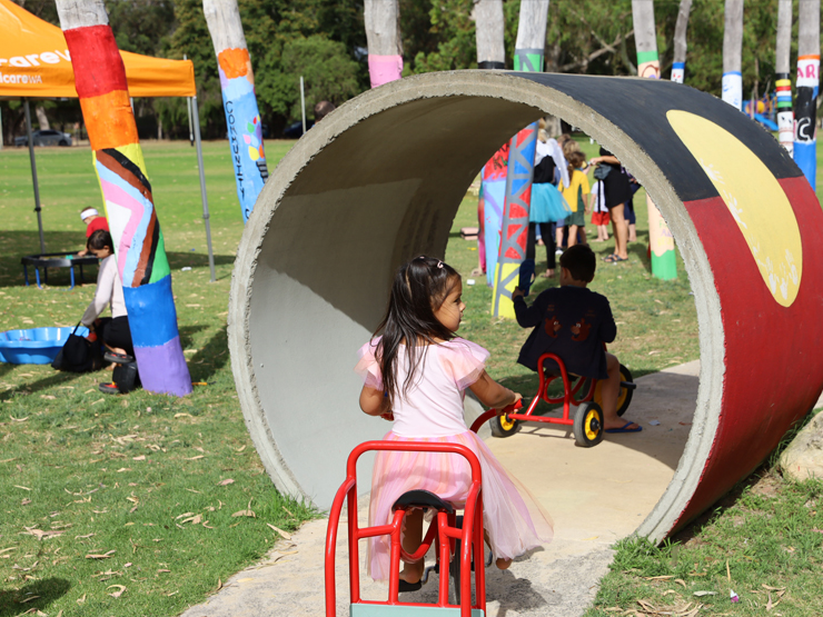 Child on bike cycling through a circular tunnel with the Aboriginal flag painted on it in a playground