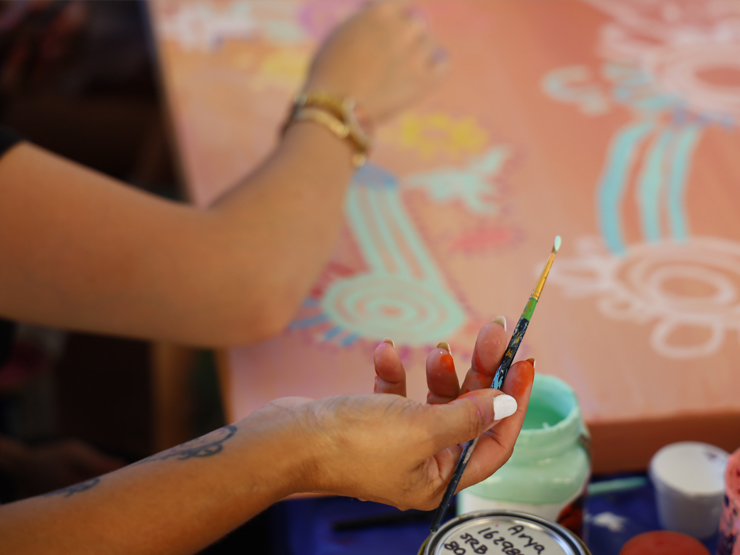 A person holding a paint brush in their hands painting on a canvas that has Aboriginal motif on it