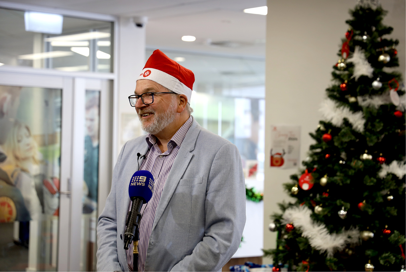 Mark Glasson CEO wearing a Santa hat standing in front of a Christmas tree speaking to media.