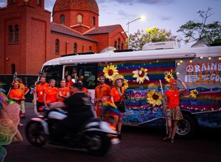Anglicare Team in the Pride Parade standing in front of a rainbow bus with giant paper sunflowers.